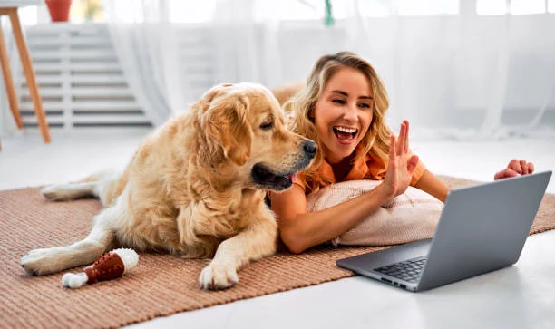 A smiling young woman lying on a woven rug next to her Golden Retriever, waving at a laptop screen during a video call in a bright indoor room.