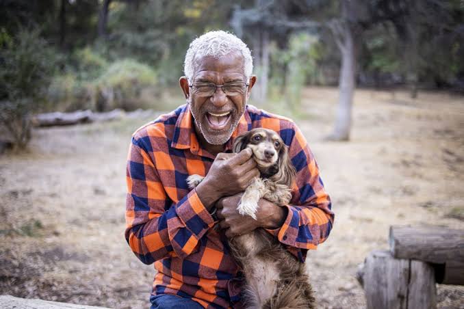A joyful older man with white hair and glasses, wearing an orange and blue plaid shirt, holding a small brown and white dog close to his chest and laughing heartily while outdoors in a wooded park setting.