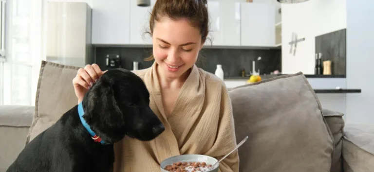 A smiling young woman wearing a beige robe sitting on a sofa, gently petting the head of a black dog with a blue collar, while holding a bowl of cereal in her lap, with a modern white kitchen visible in the background.
