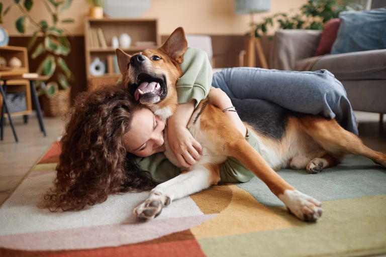 A woman with curly brown hair lying on a colorful patterned rug in a cozy living room, playfully hugging a brown, black, and white dog that is lying on its back with its mouth open in a happy, panting expression.